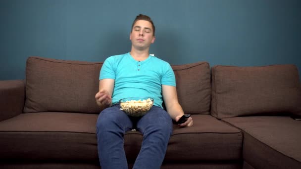 Ein junger Mann schaut fern und isst Popcorn. der Kerl fängt Popcorn mit dem Mund. auf der Couch sitzen. die Aussicht vom Fernseher. Blick in die Kamera - Filmmaterial, Video