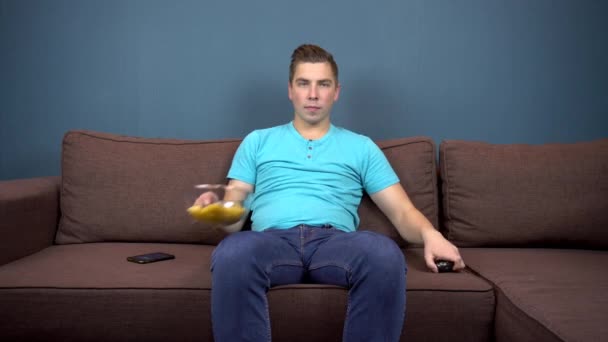 Ein junger Mann sitzt auf einem Sofa, isst Chips und sieht fern. die Aussicht vom Fernseher. Blick in die Kamera - Filmmaterial, Video