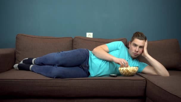 Un joven está viendo televisión y comiendo palomitas de maíz. El tipo está acostado en el sofá. La vista desde el televisor. Mirando a la cámara
 - Metraje, vídeo