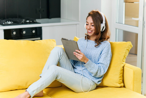 Splendid cheerful woman with brown hair and stylish clothes rests on the kitchen sofa and using laptop to listen music in headphones. - Fotografie, Obrázek