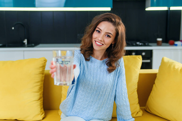 Adorable cheerful wavy-haired woman looking at camera with a glass of fresh water on the kitchen background - Фото, зображення