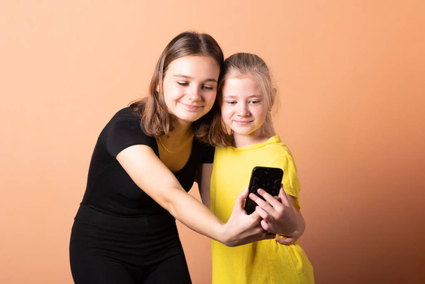 Two sisters take a selfie on a smartphone, on a light orange background. - Foto, imagen