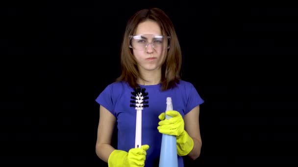 Eine junge Frau kratzt sich mit einer Klobürste am Kopf. Frau in Schutzbrille und Handschuhen mit Werkzeug für die Toilettenreinigung. Mädchen hält Klobürste und Spray. auf schwarzem Hintergrund - Filmmaterial, Video