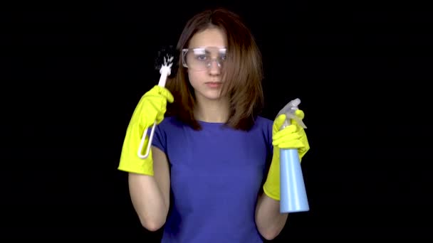 Egy fiatal nő fésüli a haját egy WC kefével. Nő biztonsági szemüvegben és kesztyű szerszámokkal a WC tisztítására. A lány WC-kefét és spray-t tart. Fekete alapon - Felvétel, videó