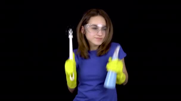 Uma jovem está dançando com uma escova de banheiro e spray. Mulher em óculos de segurança e luvas com ferramentas para limpar o banheiro. Menina segura uma escova de vaso sanitário e spray de água. Em um fundo preto
 - Filmagem, Vídeo
