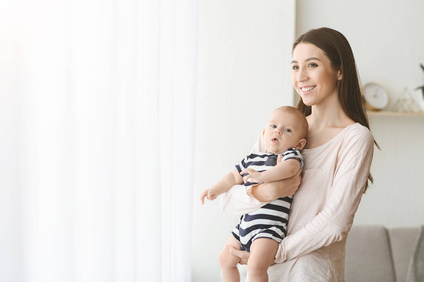 Dreamy woman with little baby on hands standing near window - Photo, Image