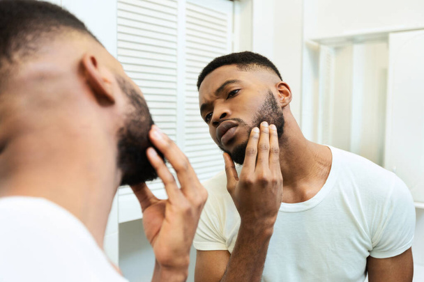 Концентрированный африканский парень, смотрящий на бороду в зеркало ванной
 - Фото, изображение