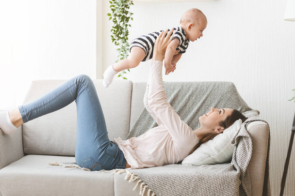 Mother lifting newborn baby in air while lying on sofa - Photo, Image