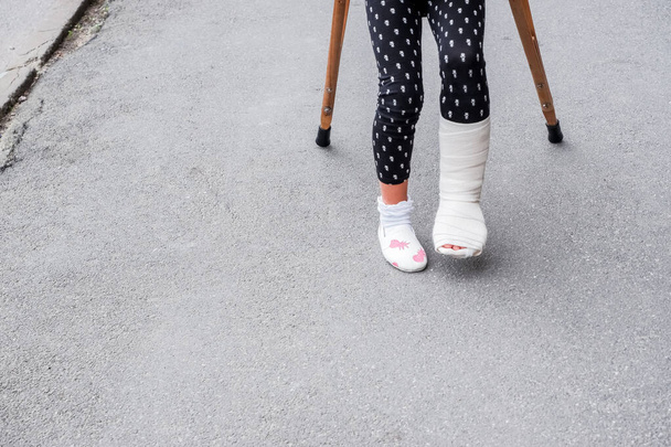 kid with broken leg is on crutches on the street. conceptual photo depicting a child with a broken leg on a holiday, on the school holiday.Girl injured in feet has bandage with crutches on asphalt - Φωτογραφία, εικόνα