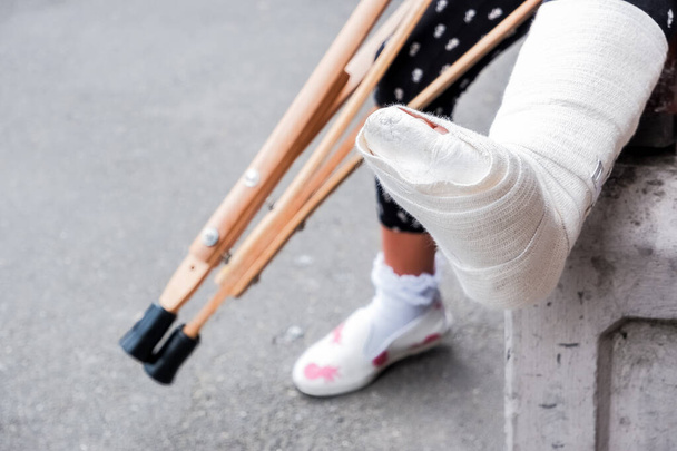 認知できない少女が壊れた足と松葉杖を持ってベンチの上の路上に座っている。松葉杖を持つ障害のある少女。トランポリンでジャンプ中の事故。足首の骨折. - 写真・画像