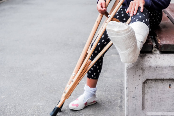 onherkenbaar meisje zit op straat op een bank met een gebroken been en krukken.gehandicapt meisje met krukken.een ongeval tijdens het springen op een trampoline. Een enkelfractuur. - Foto, afbeelding