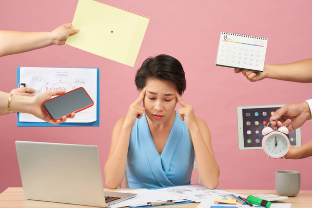 Overworked young employee refuses all things, frowns face in annoyance, sits at desktop with paper documents and notepad, isolated over pink background. Female workes bothered by many questions - Foto, Imagem