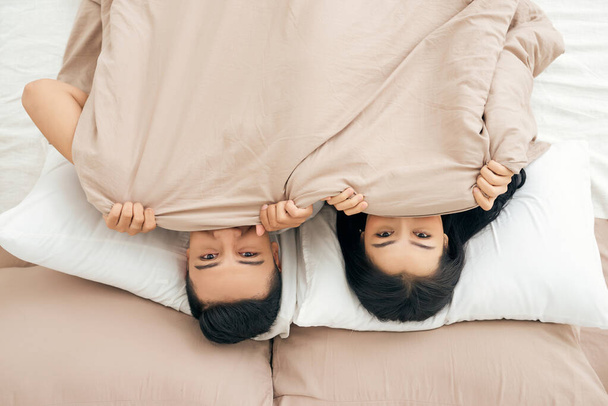 Divertida pareja casada tumbada en la cama y escondida bajo una manta blanca, mirando a la cámara con los ojos llenos de alegría
. - Foto, Imagen