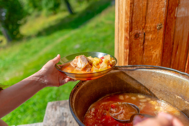 Χέρια κορίτσι μαγειρεύει σε εξωτερικούς χώρους βρίσκεται σούπα με κρέας και λαχανικά borscht από ένα μεγάλο καζάνι τηγάνι στο ξύλινο πάτωμα, σε ένα σπίτι δίπλα σε ένα γρασίδι στο δάσος, σε ένα πιάτο. - Φωτογραφία, εικόνα