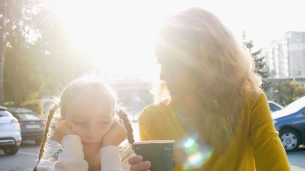 Fiatal anya sárga ingben, eldobható csésze kávé és a kis aranyos lánya beszél ül korán reggel a kávézó teraszán napkelte háttér - Felvétel, videó
