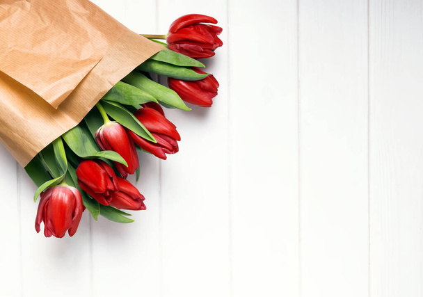 Red tulips wrapped in craft paper on the white wooden table with copy space - Фото, изображение
