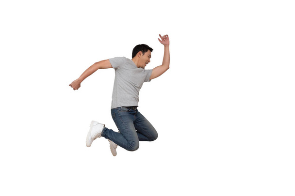 Full length portrait of an excited young Asian man in gray t-shirt jumping while celebrating success or dancing with music isolated over white background - Photo, Image