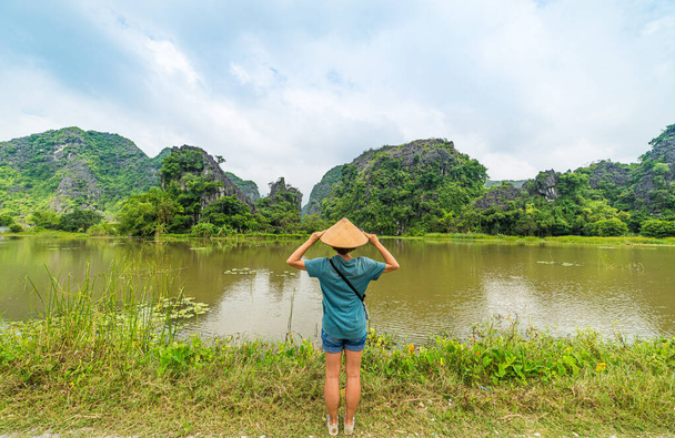 ベトナムの帽子をかぶった女性は、ベトナムのタム・コック・チャン・アン・ニンビン観光地のユニークな景色を見ています。水田の間にカルスト岩の頂点。白人女性は休暇中に楽しんでいます. - 写真・画像