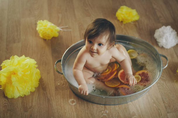 A little girl is bathing in a bath with lemon and grapefruit. A child washes in a basin among yellow flowers. concept of childhood, health care, IVF, hygiene, citrus therapy - Фото, изображение