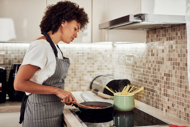 Mixed race woman in apron standing next to stove and stirring tomato sauce. On stove are saucepan and pot with spaghetti. - Photo, Image