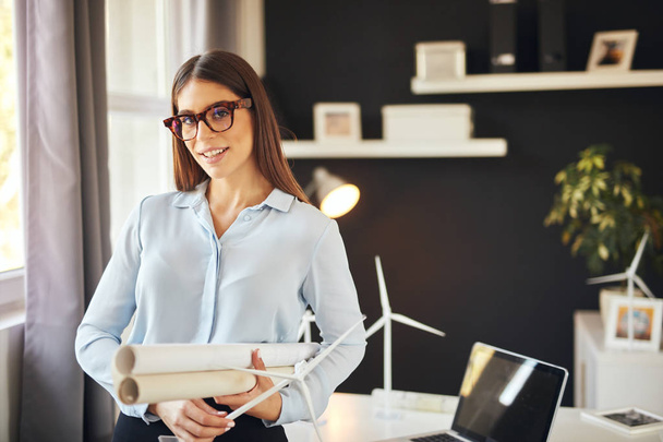 Attractive smiling businesswoman in formal wear and with eyeglasses standing in modern office and holding blueprints and windmill model. Sustainable development concept. - Zdjęcie, obraz
