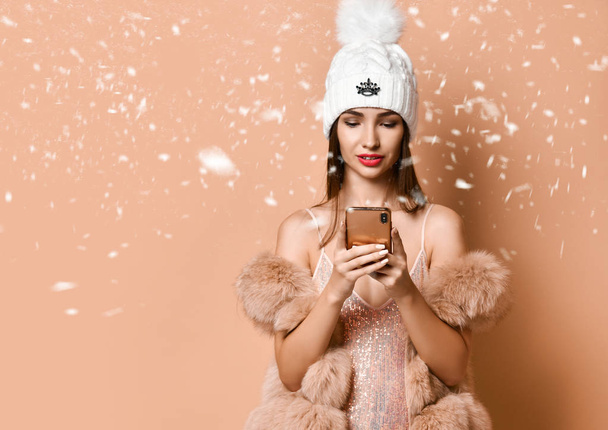 Young girl in party dress and fur vest is spending time with her smartphone, social networks under the snow on beige - Foto, Imagem