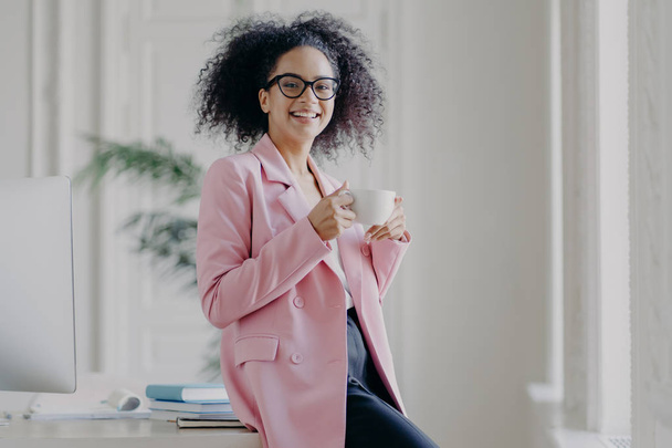 Photo of relaxed businesswoman holds cup of hot drink, has coffee break, stands near her workplace in spacious white cabinet wears spectacles long pink jacket works in office. Time for rest after work - Foto, afbeelding