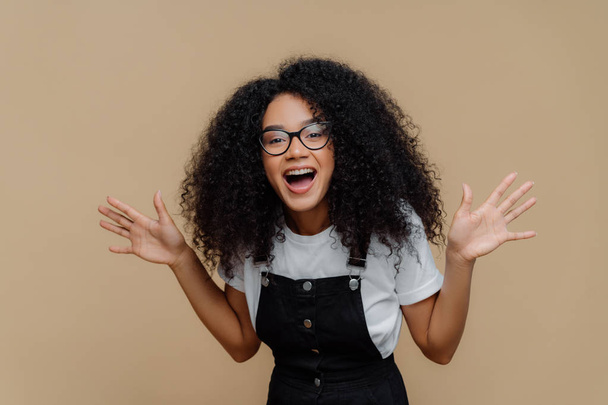 überglückliche afrikanisch-amerikanische Frau lacht, hebt Handflächen, ist gut gelaunt, trägt durchsichtige Brille, lässiges T-Shirt und Overalls, posiert vor braunem Hintergrund, fühlt Glück und Vergnügen - Foto, Bild
