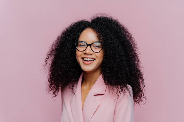 Positive curly haired businesswoman wears spectacles, formal wear, has bushy hairstyle, being in good mood after successful day at work, poses against violet background. Monochrome. Happy director - Photo, Image