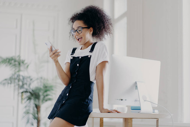 Крытый снимок счастливо удивленной молодой неопытной афроамериканской женщины носит очки, белую футболку и сарафан, использует мобильный телефон для чата в Интернете, позирует рядом с рабочим столом с компьютером
. - Фото, изображение