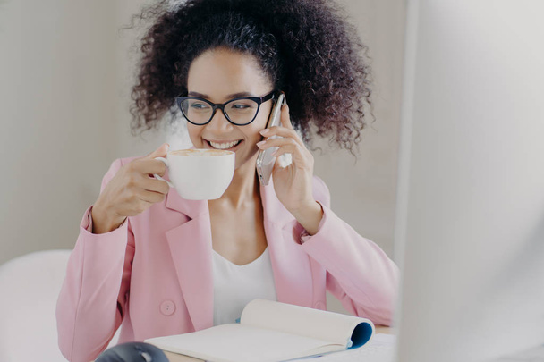 Cropped image of happy young African American woman makes phone call, drinks aromatic latte or espresso, poses in office interior, has pleasant smile wears optical glasses and rosy formal jacket - Photo, Image
