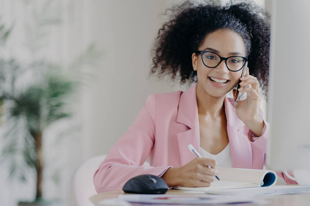 Happy African American woman with curly hairstyle, writes some information, speaks via cell phone, sits at desk against blurred office background. People, technology, communication, business concept - Foto, Imagen
