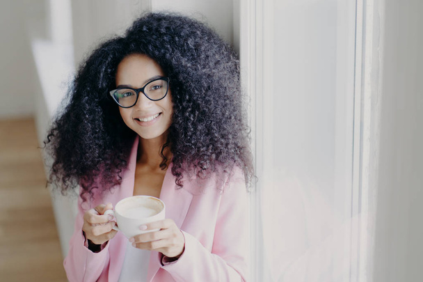 Photo of pleasant looking curly haired lady with toothy smile, dressed formally, poses indoor, holds mug of latte, has coffee break after long hours office work, looks through eyewear, feels happy - Photo, Image