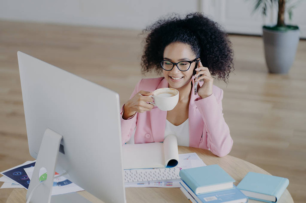 Glad dark skinned business lady looks happily at computer, drinks fresh hot beverage, holds modern mobile phone, dressed elegantly, poses against office interior, sits at desk with textbook, notepad - Foto, imagen