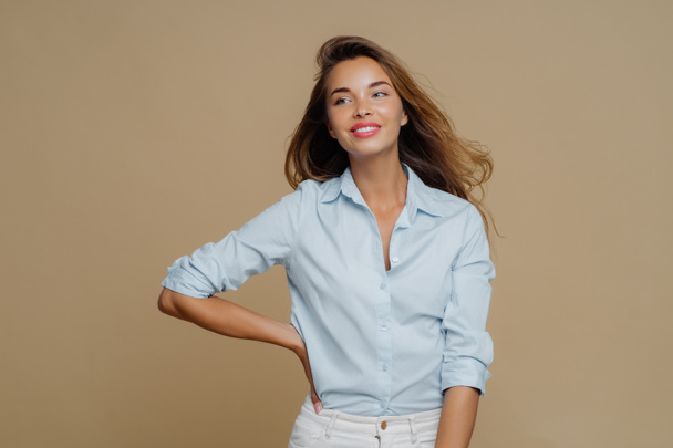 Shot of pretty cheerful European woman keeps one hand on waist, looks aside, has charming smile wears shirt and trousers, has slim perfect figure, enjoys shopping day, isolated over brown background - Foto, Imagem