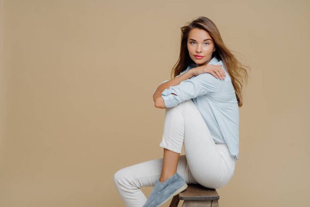 Studio shot of restful beautiful Caucasian woman sits on chair, wears shirt, white trousers and shoes, keeps hands crossed over body, has confident look at camera, isolated over brown background. - Photo, Image
