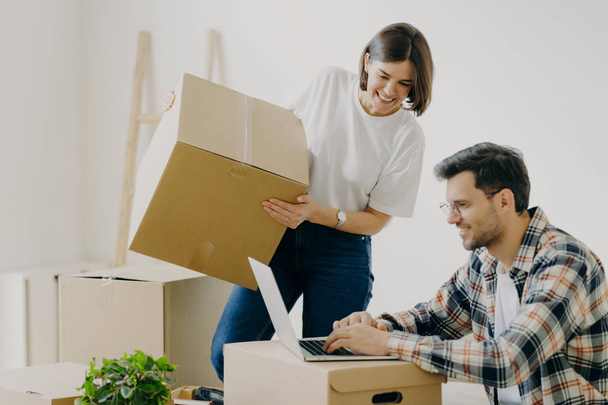 Freshly married family couple pose in their new apartment, unpack boxes with belongings, glad man sits in front of laptop computer, searches ideas for designing, have domestic routine problems - Foto, immagini