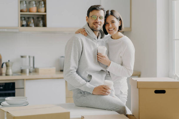 Image of glad family couple hug and stand closely to each other, drink takeout coffee, look with smile at camera, dressed in casual clothes, surrounded with cardboard boxes, spend free time in kitchen - Photo, Image