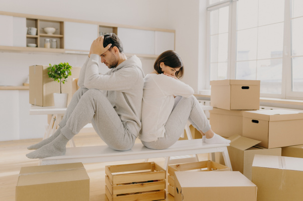 Unhappy young married woman and man have to leave house, move in other place, sit back to each other pose in empty room with stack of boxes, wear domestic clothes and socks, have some problems - Photo, Image