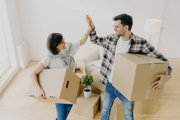 New home. Happy woman and man celebrate moving to new apartment, pose in empty room with cardboard boxes and couch in background, give each other high five. People, home, real estate concept - Foto, immagini