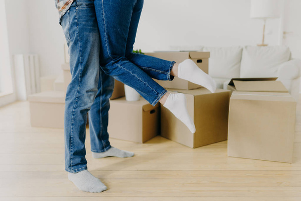 Cropped shot of caring man lifts his wife up, wears jeans and socks, just moved in new home, pose around cardboard boxes on floor, excited about moving. Unrecognizable couple relocate in new place - Photo, Image