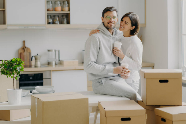 Caring woman embraces husband with love, drink takeaway coffee, pose in modern kitchen with unpacked boxes around, move into new apartment for living, rent flat, unpack belongings, have break - Photo, Image