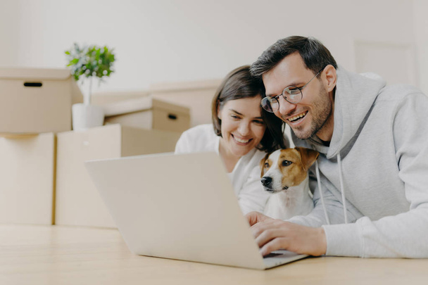 Overjoyed family couple laugh as watch online stores on modern laptop computer with their dog, have break after unpacking carton boxes, home renovation, being in good mood. Newly rented house. - Photo, Image