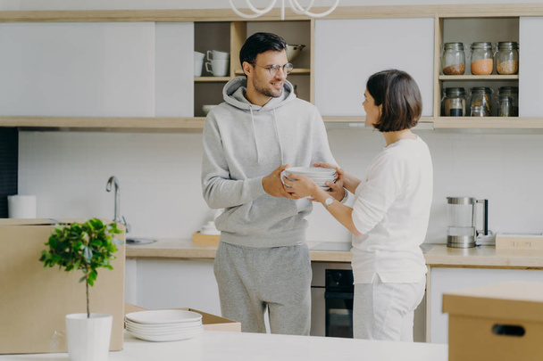 Zdjęcie szczęśliwego męża i żony rozpakować rzeczy po wprowadzeniu się do nowego domu, nosić białe talerze, pozować do wnętrza kuchni, bawić się, ubrany w ubrania domowe. Ludzie i koncepcja ruchu - Zdjęcie, obraz