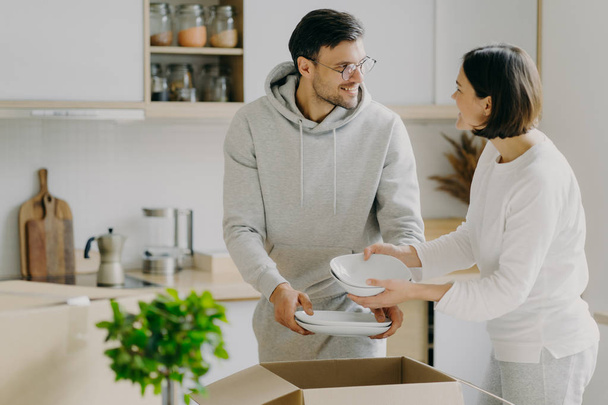 Delighted housewife and her husband unpack cardboard boxes with new tableware, carry plates, move into new apartment, look gladfully at each other, pose agaist kitchen interior. Relocation concept - Photo, Image
