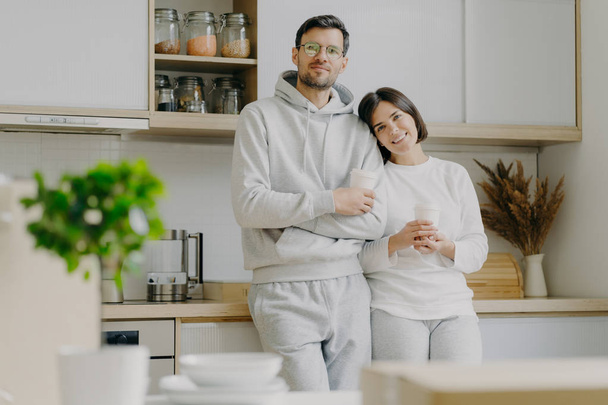 Shot of relaxed family couple stand in kitchen, drink takeaway coffee, wear casual outfit, pose in kitchen interior, bought new apartment for living. People, real estate and mortgage concept - Fotografie, Obrázek
