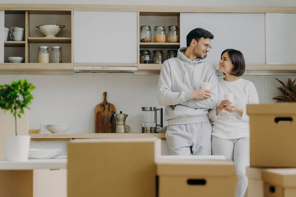 Indoor shot of lovely newlywed couple talk to each other with pleased expressions, drink takeaway coffee from paper cups, pose in modern kitchen with new bought furniture, change place of living - Photo, Image