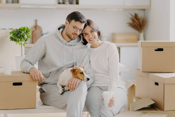 Tired married family couple drink aromatic takeaway coffee, sit closely to each other, pose with pedigree dog near big carton boxes, pose against kitchen interior, move into new modern apartment - Photo, Image