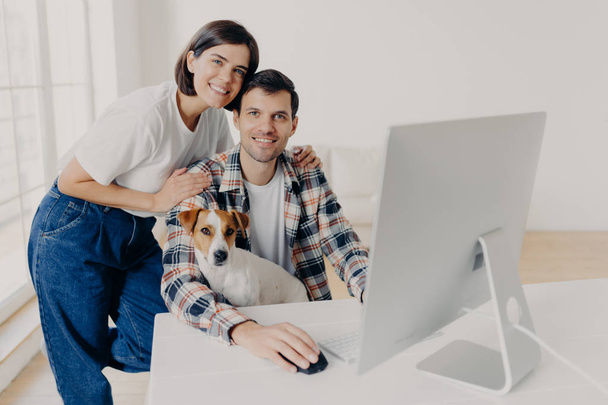 Photo of male freelancer works remotely at computer with dog, his affectionate wife embraces and helps with some questions, pose in big room at workplace, discuss some issues and family budget - Φωτογραφία, εικόνα