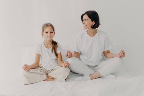 Horizontal shot of happy relaxed brunette mother and daughter pose in lotus pose, try to relax, keep legs crossed, dressed in casual white clothes, meditate on bed, pose against light background - Fotó, kép